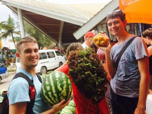 Caucasian young men smiling and carrying a watermelon and oranges at the market.