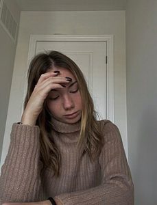 Woman sits with head down and eyes closed. Her Hand is rested on her head. Her face is sad.