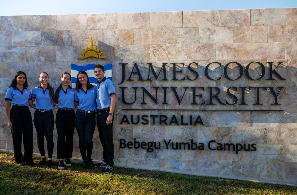 The five student authors of this education resource ebook are shown in a photo beside the entrance gate and logo signage of the James Cook University Townsville, Bebegu Yumba Campus