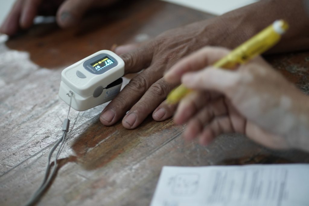 A close-up photo of a hand wearing the pulse oximeter. In the foreground, a second blurred hand is recording the readings on a piece of paper.