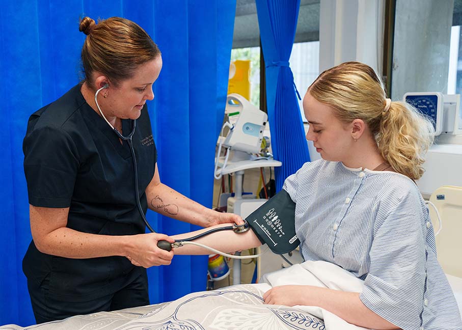 A photo of a nurse using manual methods to take a patient’s blood pressure. The patient is seated in bed while watching the standing nurse work.