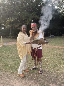 A woman and man stand together, smiling and holding a bark containing containing smoking eucalyptus leaves