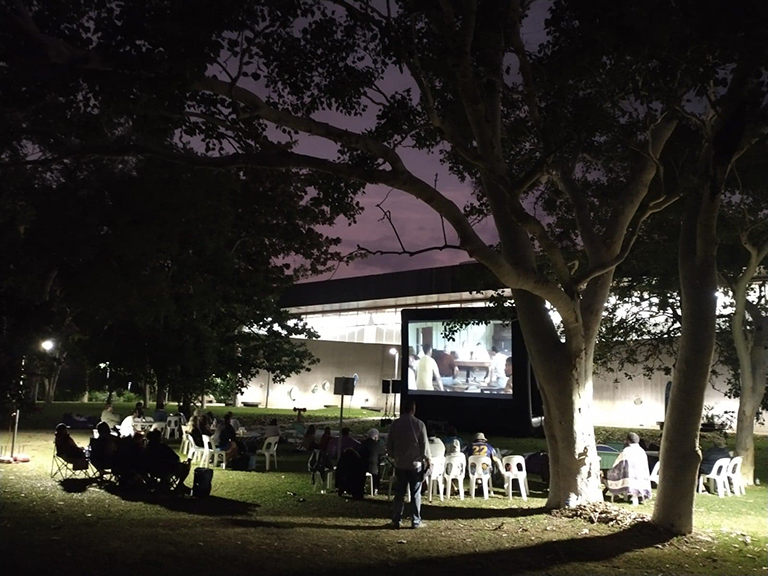 A view through the trees of an audience seated in front of a movie screen, with the Library behind.